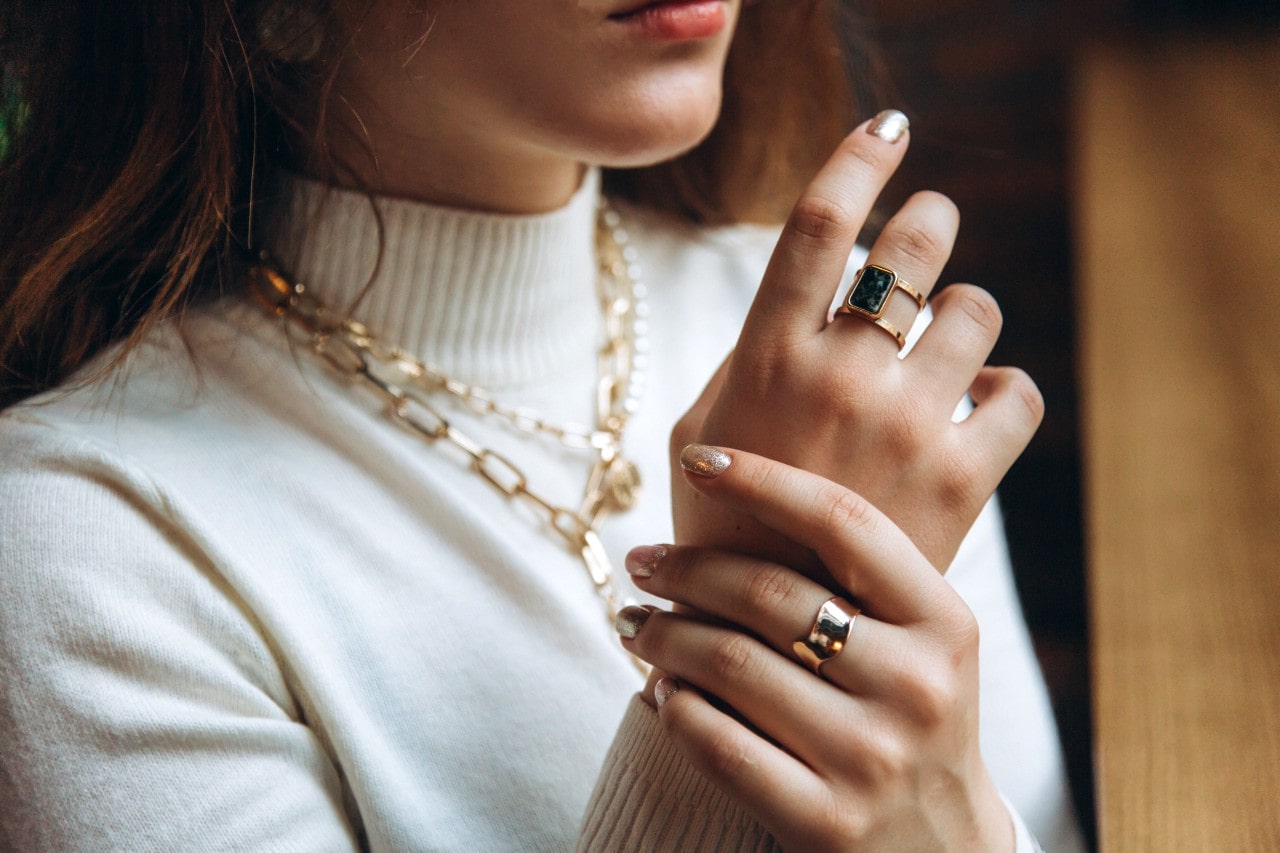 A close up of a woman in a white turtleneck, showcasing yellow gold chain necklaces and chunky rings