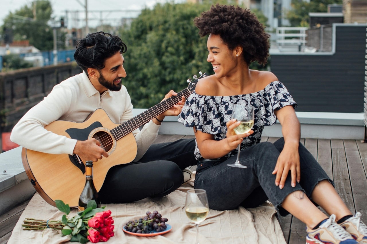 A man plays guitar for a smiling woman on a rooftop, surrounded by wine, roses, and grapes.