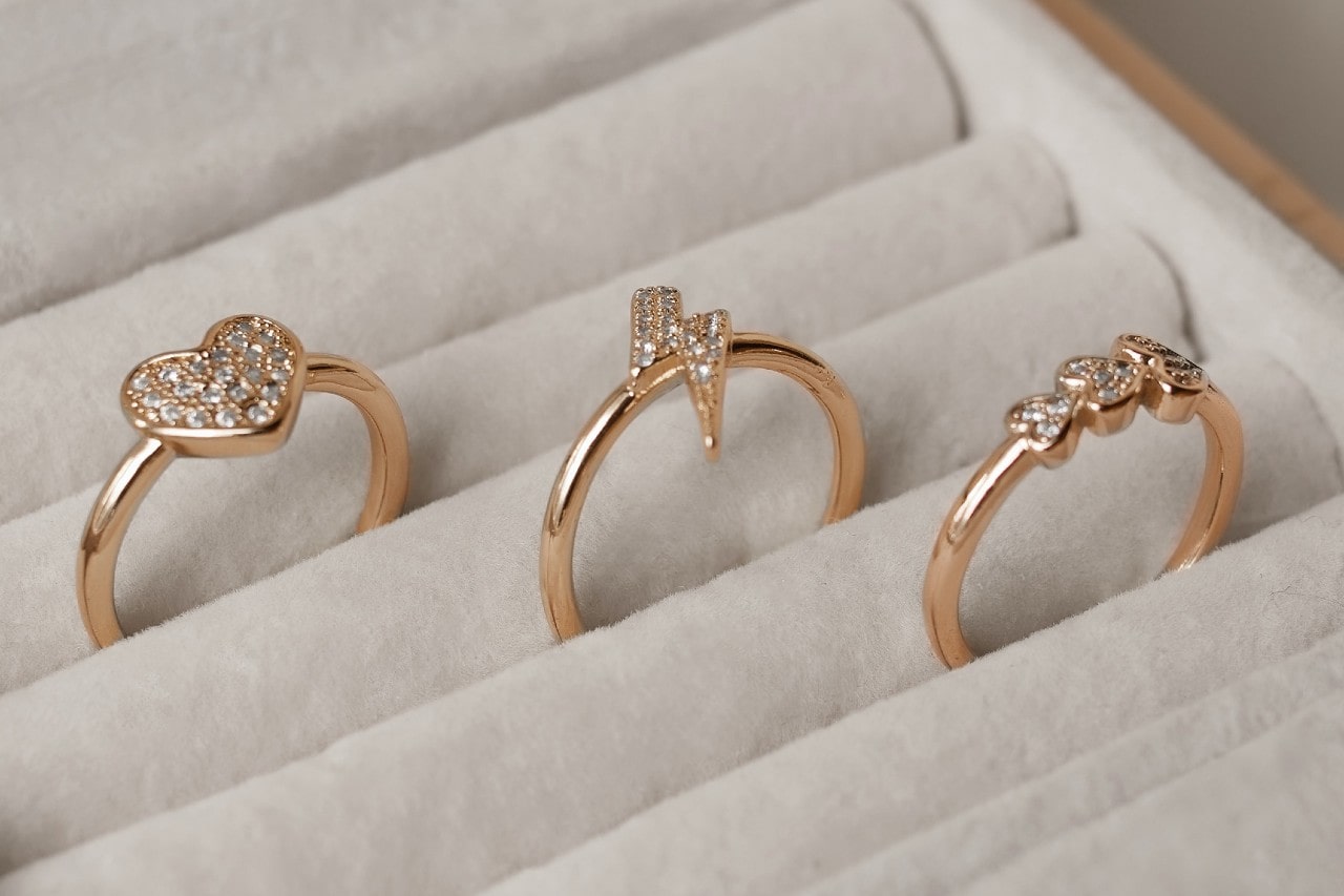 A close-up of three whimsical stackable rings, one with a large heart, another with a lightning bolt, and the last with three small hearts, displayed in an open jewelry box.