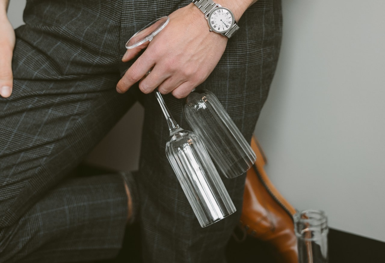 A close-up of a well-dressed man wearing an elegant watch leaning on a wall as he holds two champagne glasses.