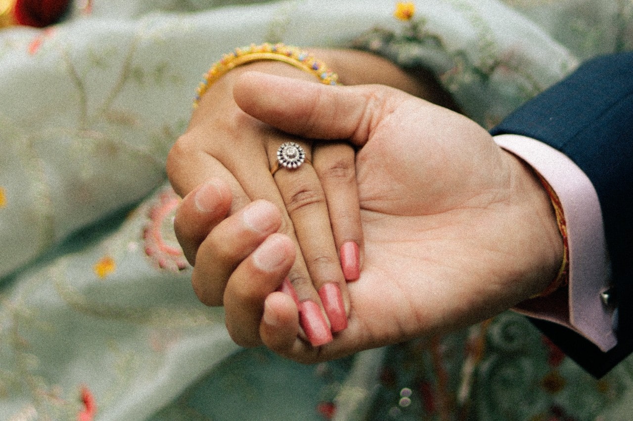 a man holds a woman&rsquo;s hand, showing off her halo engagement ring.