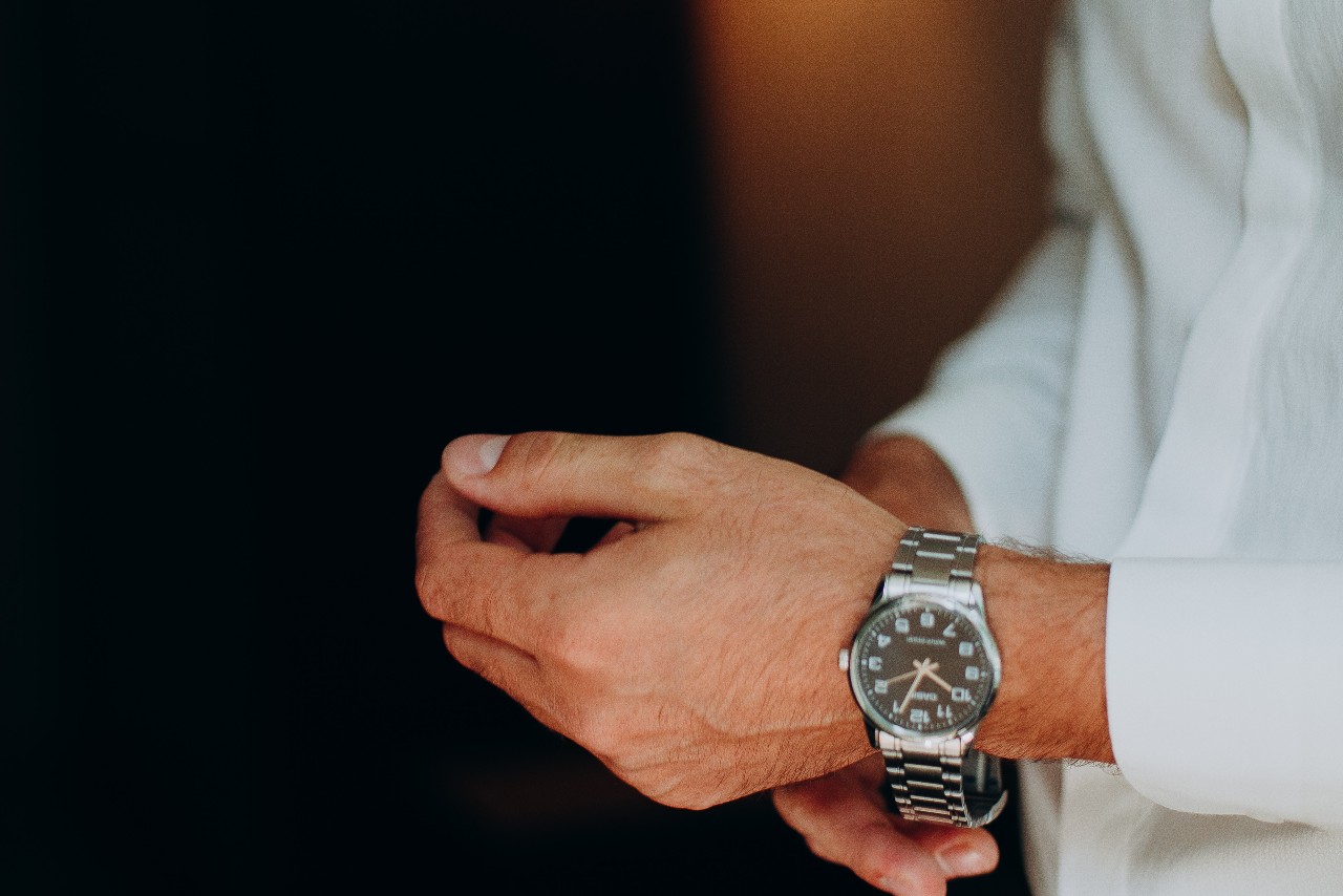 A close-up of a man in white shirt putting his stainless steel watch on his wrist against a dark background.