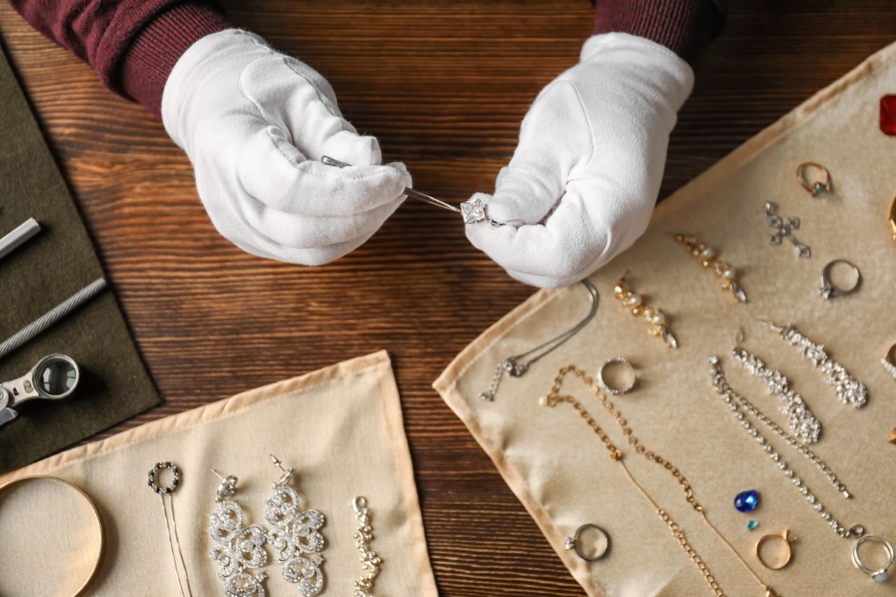 Close-up of a jeweler wearing white gloves inspecting a diamond pendant with tweezers, surrounded by an assortment of jewelry displayed on beige cloths over a wooden table.
