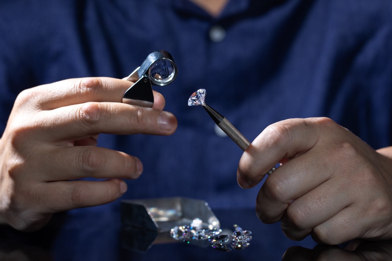 Close-up of a jeweler in a dark blue shirt examining a heart-shaped diamond with a loupe, holding it with tweezers, with loose diamonds on a black surface in front.