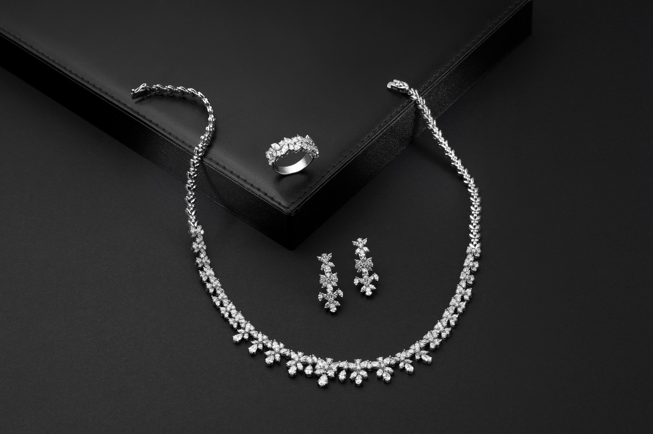 A stunning diamond necklace, ring, and matching earrings in a floral motif, glistening on a black leather display against a black backdrop.
