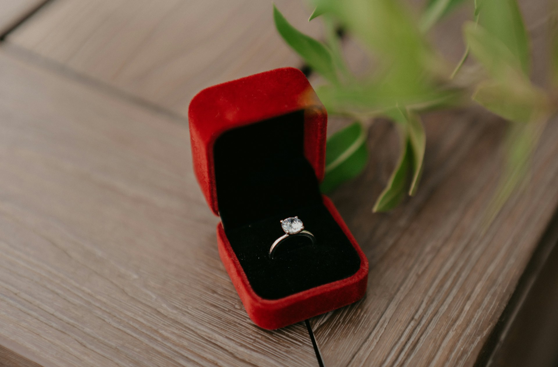 A solitaire platinum diamond engagement ring in a red velvet box placed on a wooden surface
