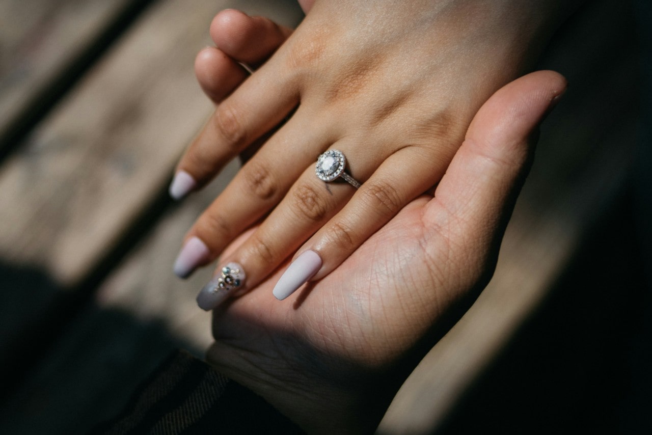 A woman’s hand with long nails showcase white gold halo diamond engagement ring, while being on top of man’s hand