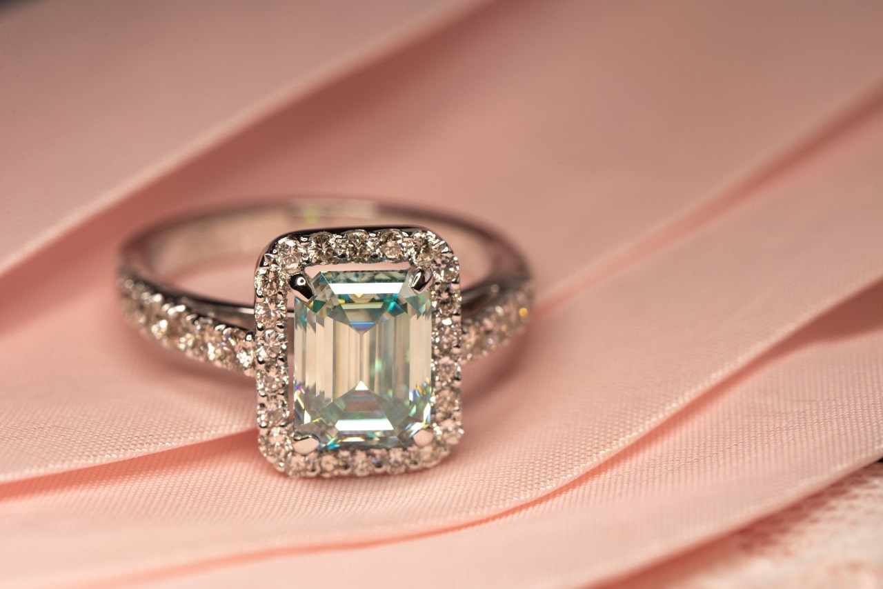 A close-up of an emerald-cut diamond ring with a halo setting and diamond-encrusted band, resting on pink fabric.