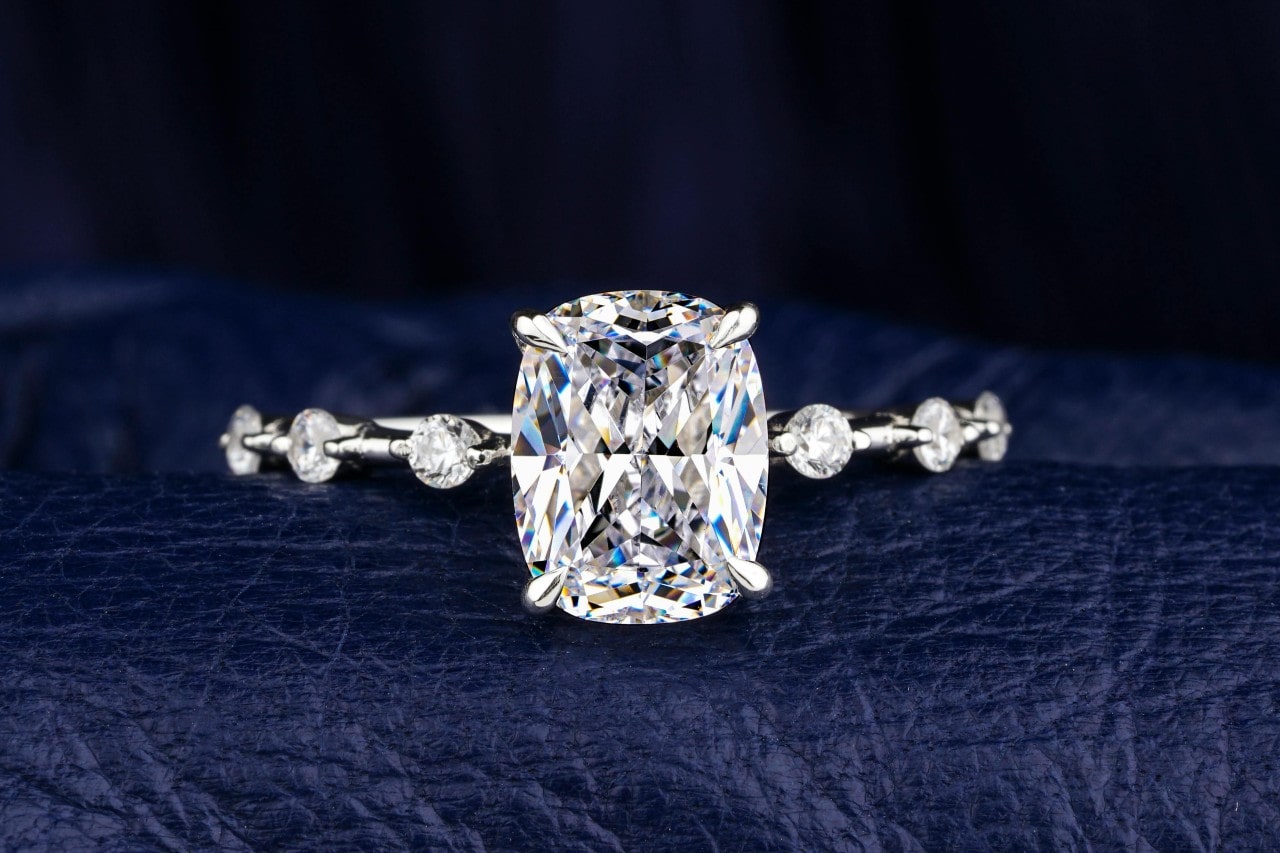 A close up of cushion cut white gold side stone engagement ring on a textured dark blue surface