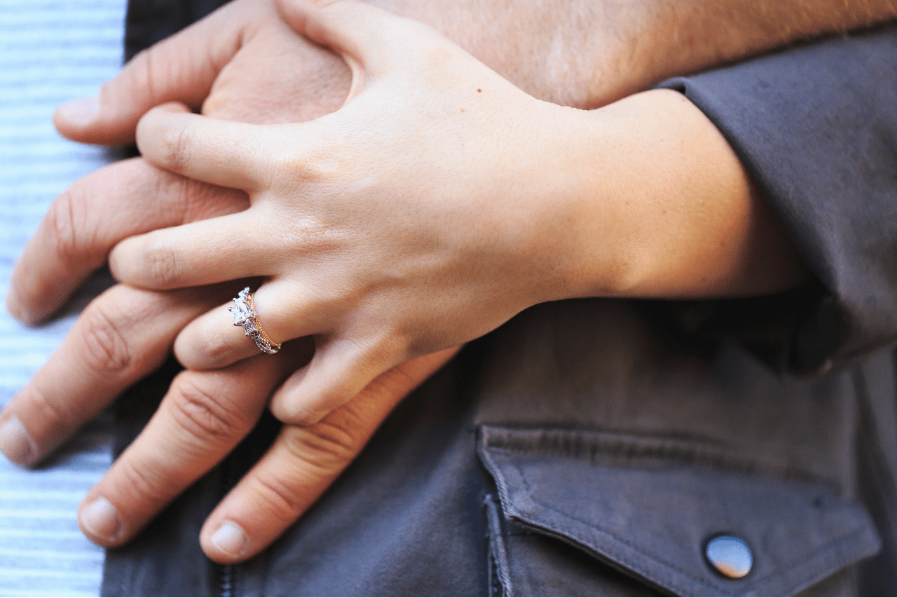 Close-up of a couple’s hands, the woman’s engagement ring resting elegantly atop the man’s hand over a gray denim jacket.