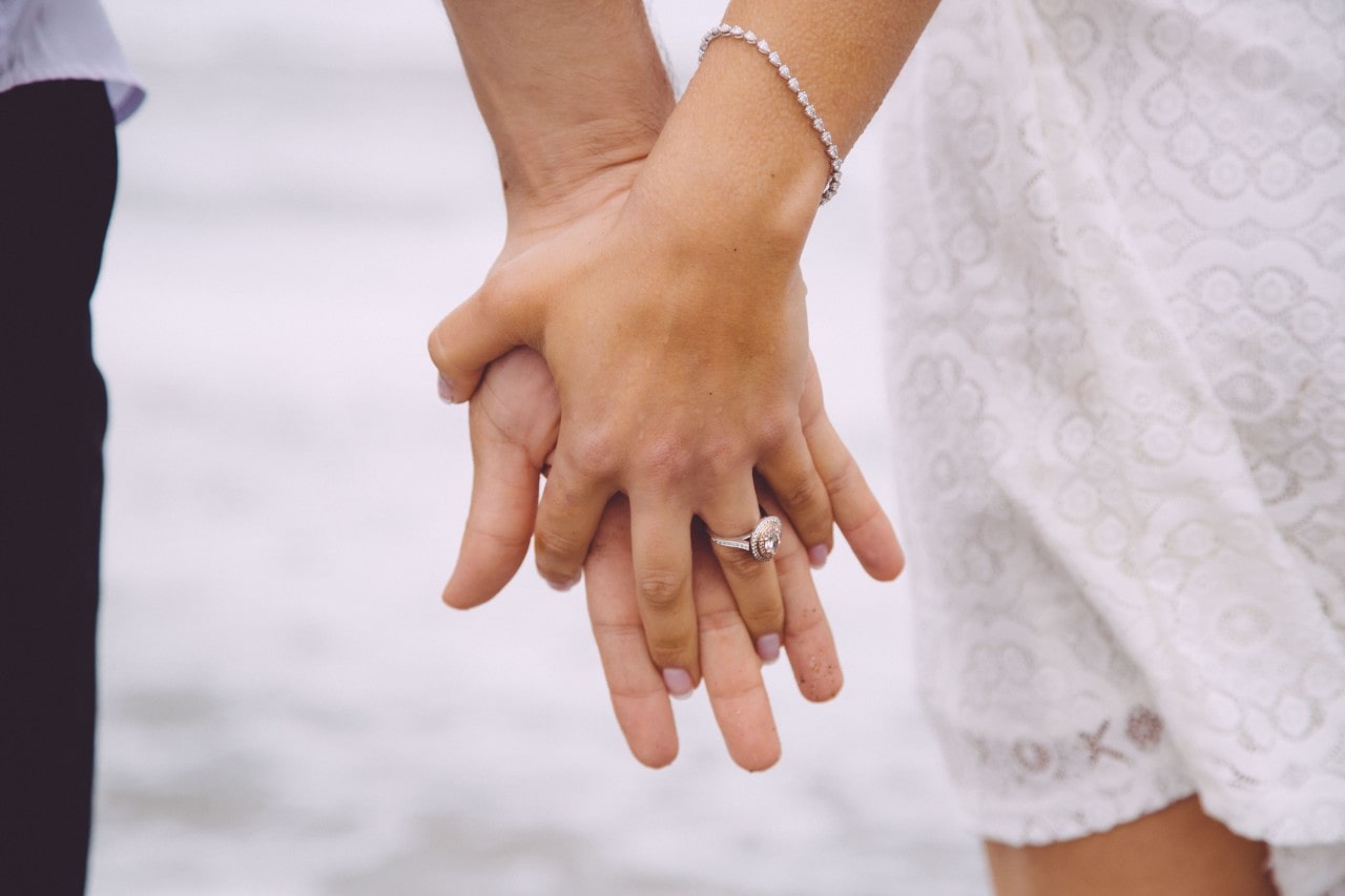 Romantic beach scene with a couple holding hands, highlighting the woman’s dazzling engagement ring.