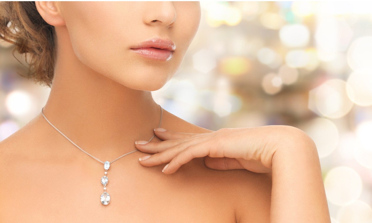 A woman with glossy lips gently touches a diamond pendant necklace against a softly blurred background.