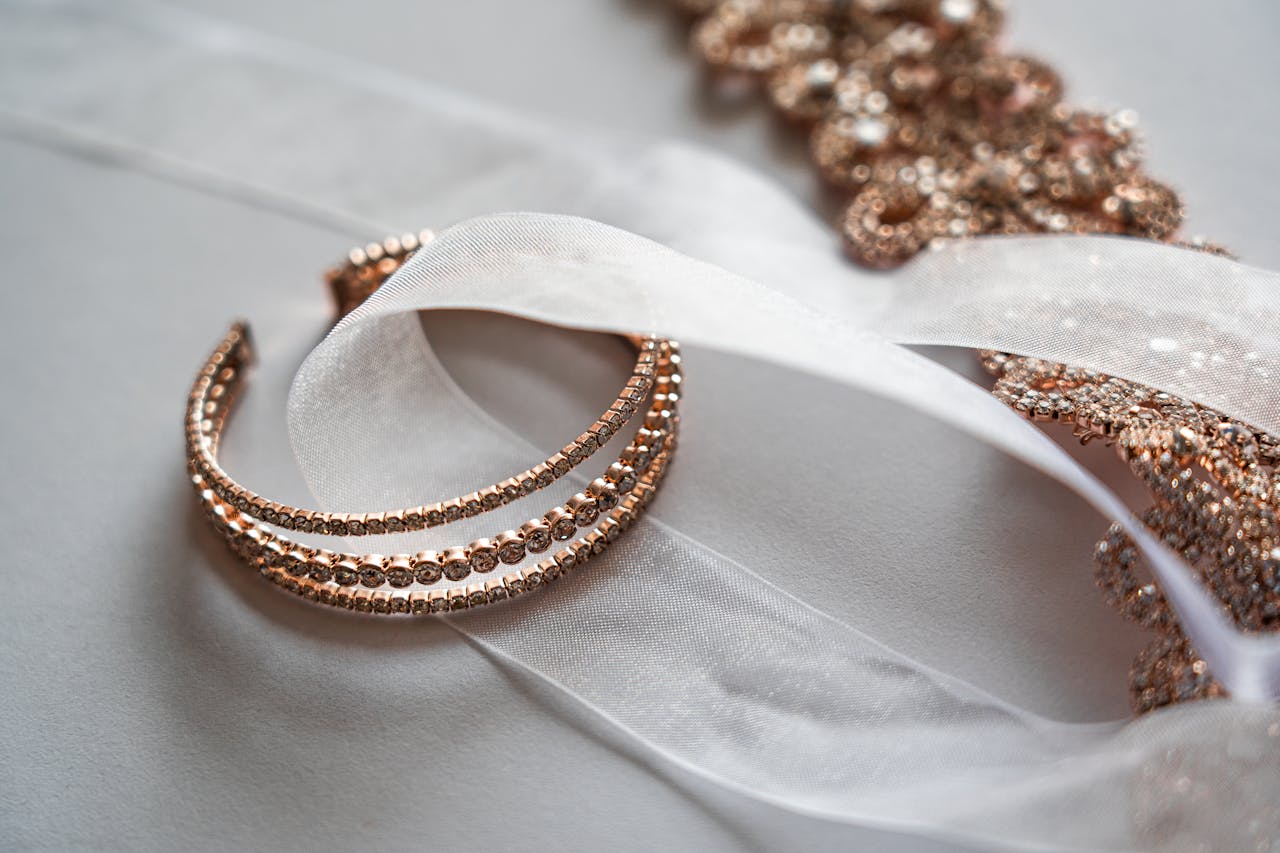 A delicate rose gold bracelet intertwined with sheer white ribbon lies on a smooth surface, with rose gold jewelry in the background.