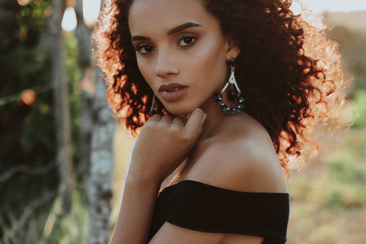 A woman in a black off-shoulder top with curly hair and dangling earrings posing against a blurred natural background.