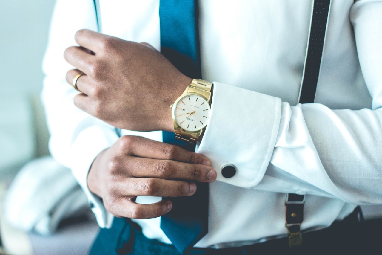 A man in white shirt fixes his cufflink, showcasing yellow gold wristwatch and polished wedding band