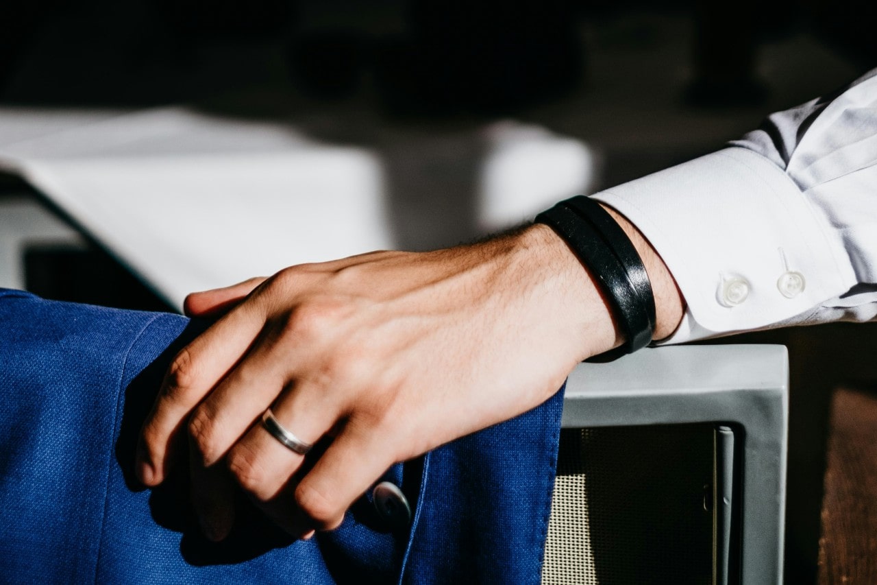 A close up of a man’s hand, showcasing white gold wedding band and rubber black bracelet.