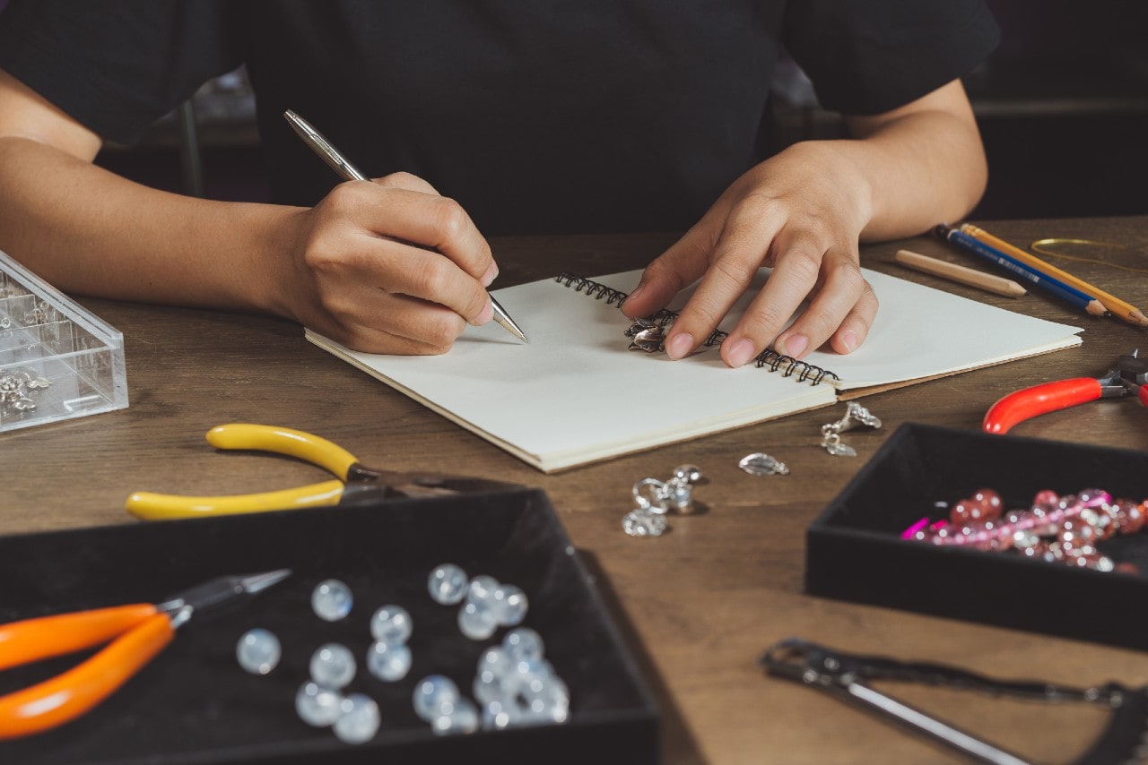 A jewelry designer sketches a piece by hand in a notebook surrounded by beads, tools, and gemstones. A jewelry designer sketches a piece by hand in a notebook surrounded by beads, tools, and gemstones.