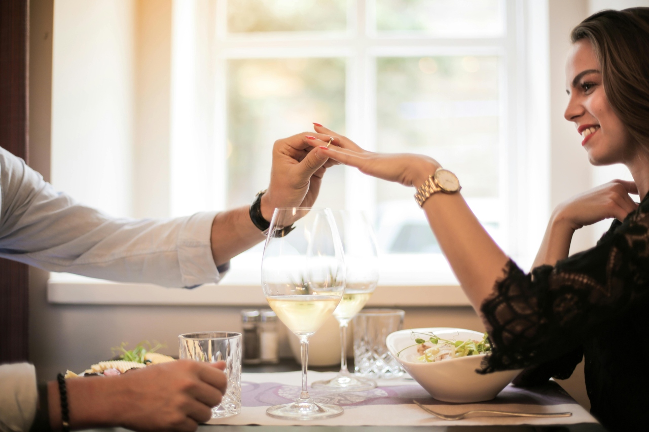A man putting a diamond ring on a woman’s finger across the dinner table, set with salads and two glasses of white wine.